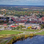 “Aerial view of downtown Bangor, Maine, showing the Penobscot River in the foreground, historic brick buildings, green parks, and surrounding autumn forests under a partly cloudy sky