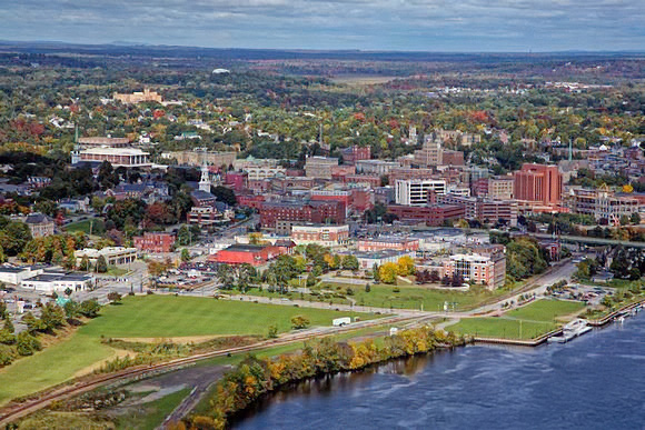 “Aerial view of downtown Bangor, Maine, showing the Penobscot River in the foreground, historic brick buildings, green parks, and surrounding autumn forests under a partly cloudy sky